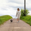 havaneser.shop Umhängeleine Klassik aus braunem Fettleder, getragen von einer Frau mit Hund auf einem Feldweg.